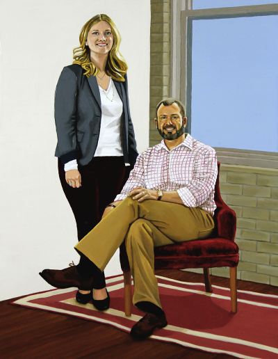 A couple pose for their portrait in a painter's studio. Jon is sitting in a red crushed velvet chair. He is wearing khaki trousers and a checkered shirt. Jen is standing. She is wearing a gray jacket over a white shirt with dark red pants.